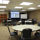 Room 235 is set round table seating. Lights are on. Projector screen is down utilizing the ceiling mounted projector. White board and lectern are up front. Air wall is up. View is from the entry door side.