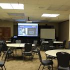 Room 235 is set round table seating. Lights are on. Projector screen is down utilizing the ceiling mounted projector. White board and lectern are up front. Air wall is up. View is from center of room.