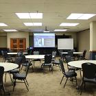Room 235 is set round table seating. Lights are on. Projector screen is down utilizing the ceiling mounted projector. White board and lectern are up front. Air wall is up. View is from the rear.