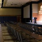Classroom style seating. Lights are dimmed. Montana State University “GOBO” is projected onto the wood panel wall. Projector screen is down. A lectern and microphone are on the former stage. Pipe and drape is set up on the far side of the room. View is from a set of entry doors.