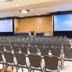 Theater style seating. Lights are all the way on. Projector screens are down. A lectern and panel tables with skirting are on the former stage. View is from the back of the room.