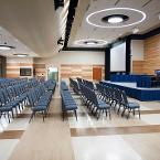 Theater style seating. Lights are all the way on. Projector screen is down. A lectern and panel tables with skirting are on the former stage. View is from a set of entry doors.