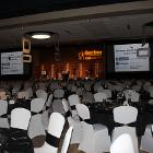 Image of decorated round tables with projector screens and stage in background.