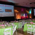 Image of tables in green tablecloths with white chairs. Screens and stage is in the background.