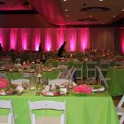 Image of squre table with green tablegloth and white chairs with staff setting up for event in the background. Walls are lit with magenta floor lights.