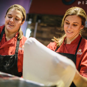 two female culinary services staff members stretching pizza dough