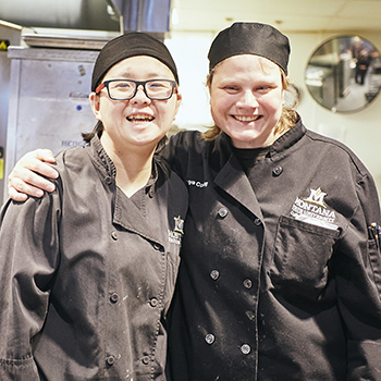 two student workers smiling while working in the dining hall kitchen