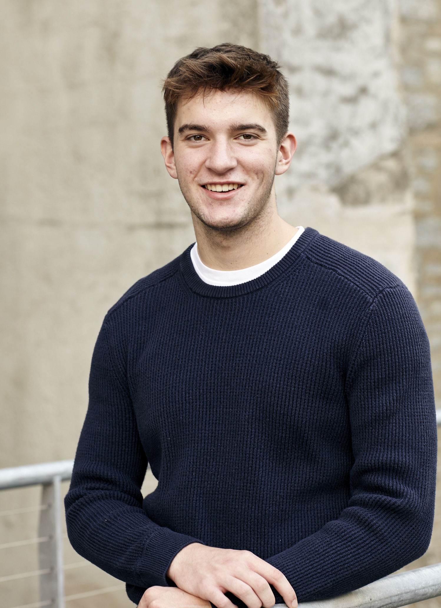 young man with dark hair and brown eyes wearing a blue swearter posing in front of a concrete wall