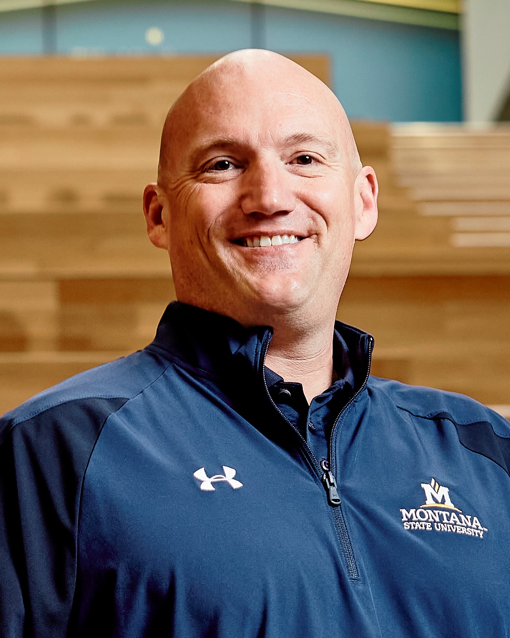 photo of man with a bald head and brown eyes wearing a blue Montana State University logo shirt, standing in front of wooden steps