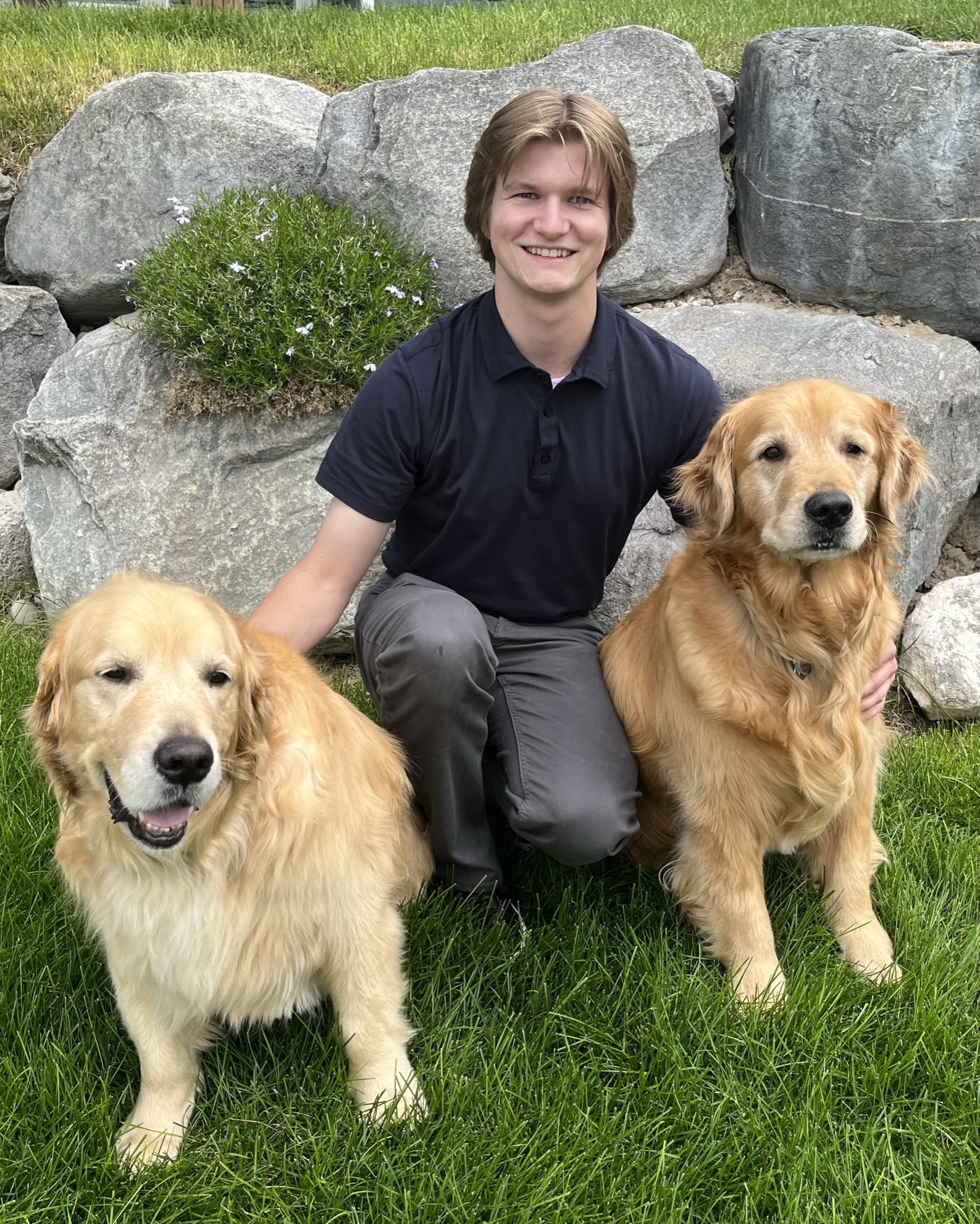 Young male with slightly longer light brown hair, blue short sleeve shirt and gray slacks, with two golden retriever dogs in front of large gray rocks and grass