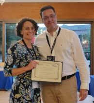 Photo of a man and a woman standing in a business lobby. The woman has dark, curly hair and is holding a certificate for the award she won.