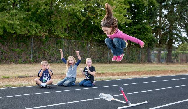 Three children sitting on a black track, one child jumping over a small hurdle.