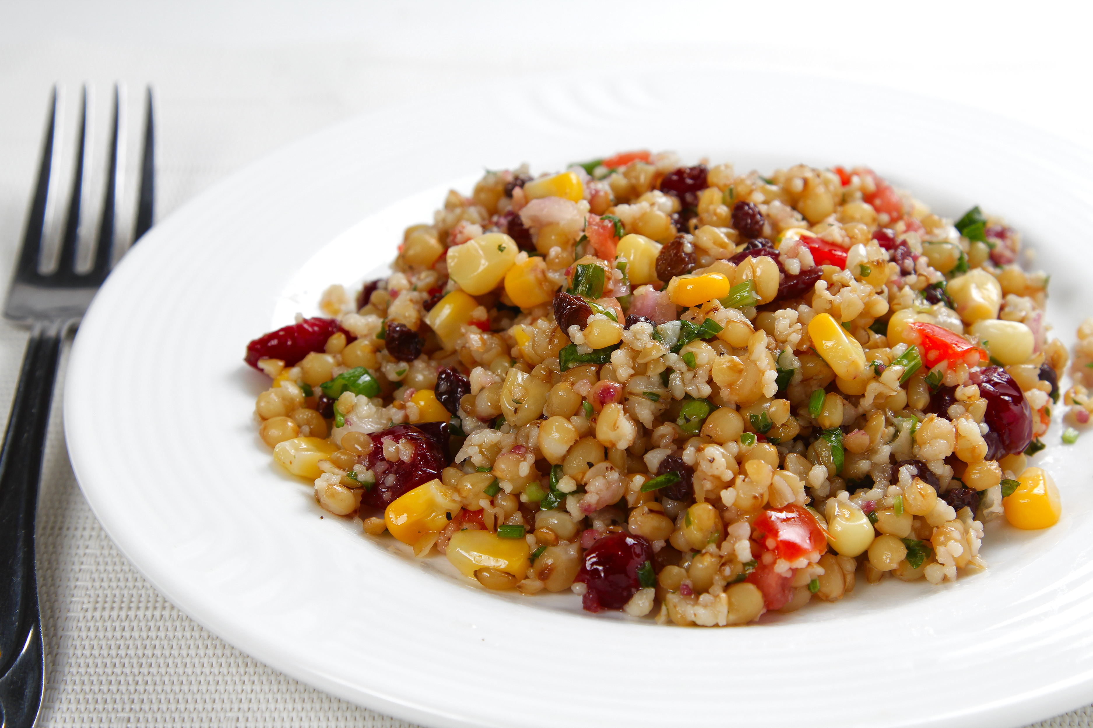 A white plate next to a fork containing barley, corn, and cranberry salad.