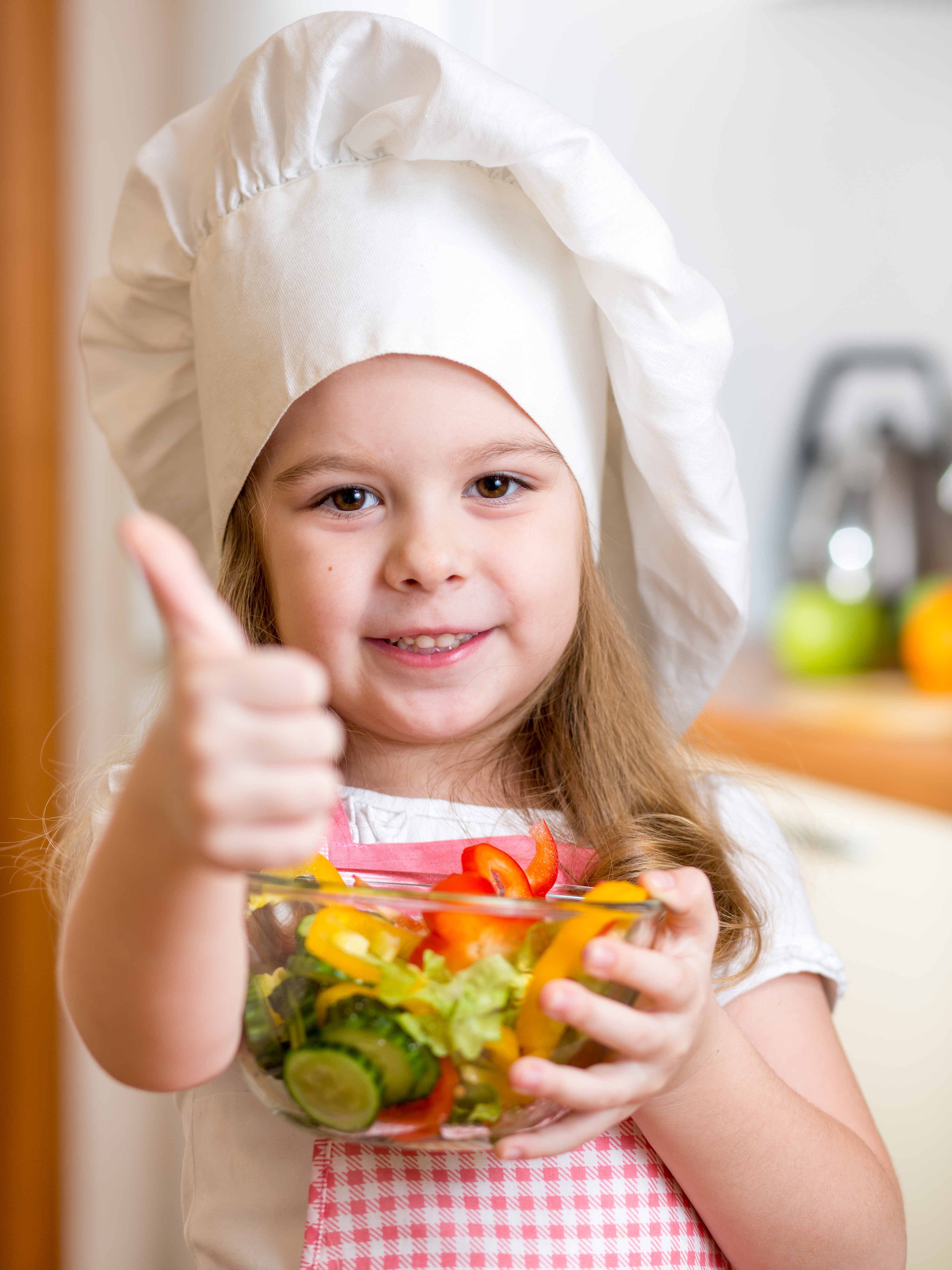 A young girl wearing a white chefs hat while holding a bowl of vegetables and giving a 'thumbs-up' sign.