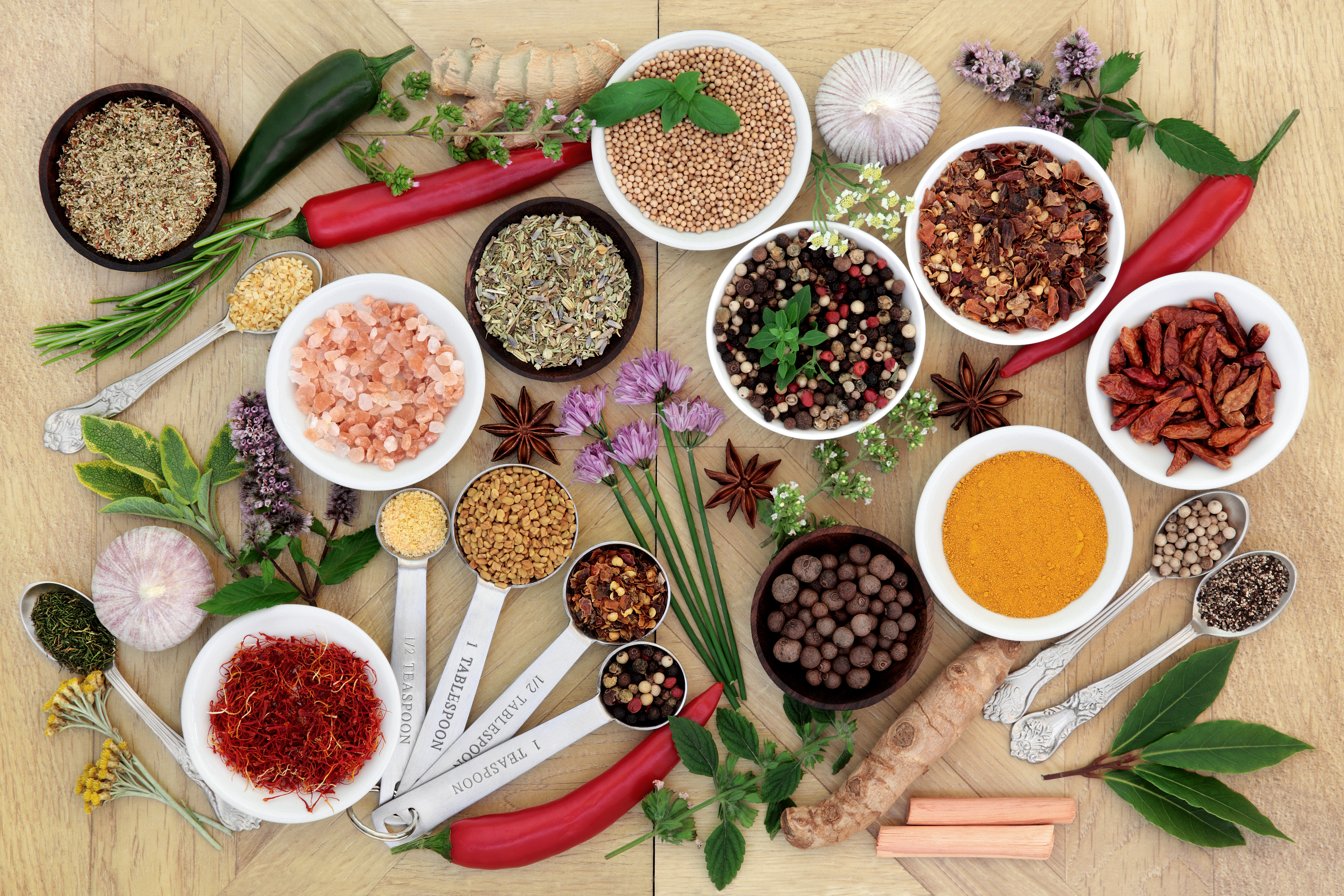 A table with bowls of seasonings and spices alongside various green herbs, red peppers, roots, and flowers.