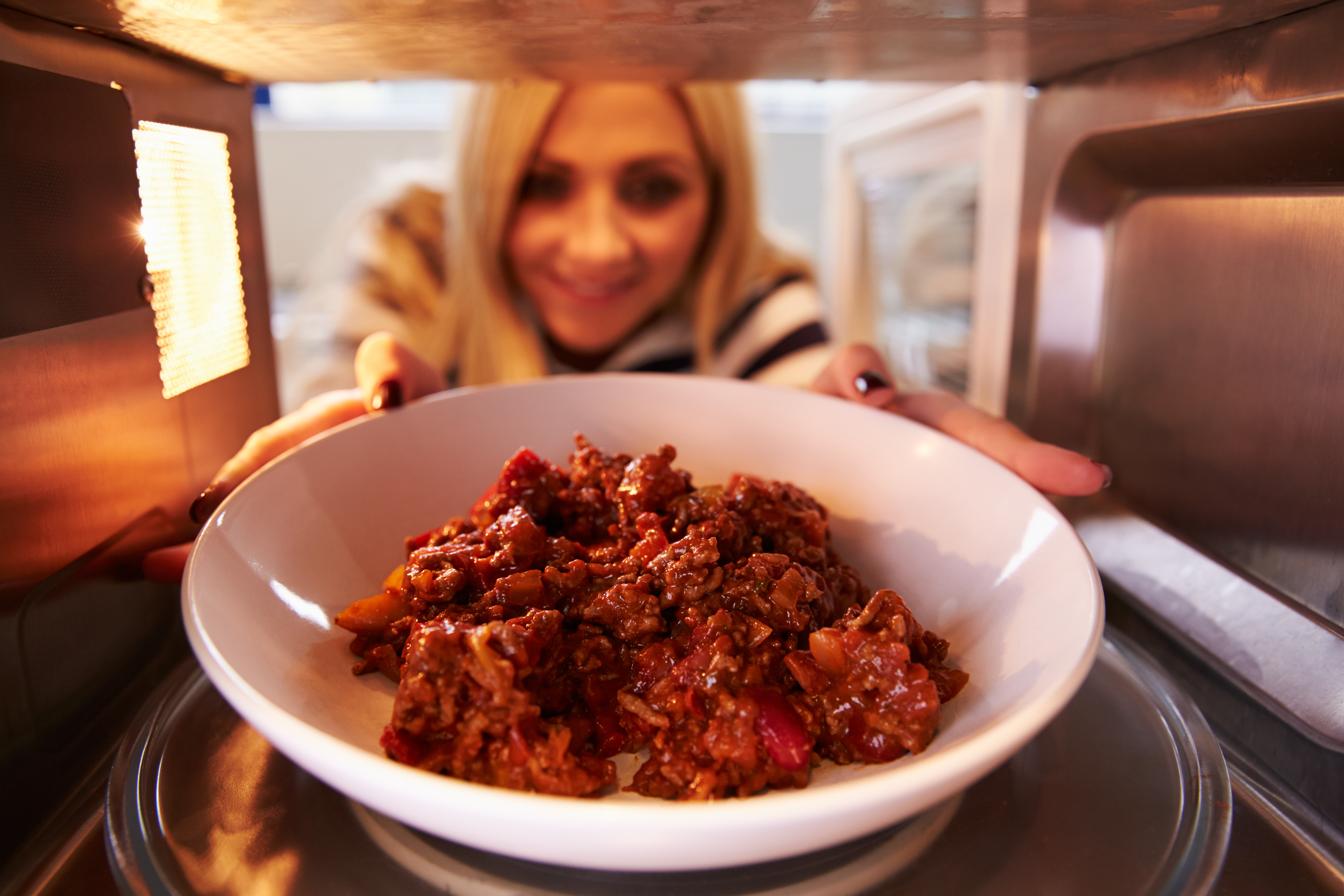 A woman place a white bowl filled with meat into a microwave.