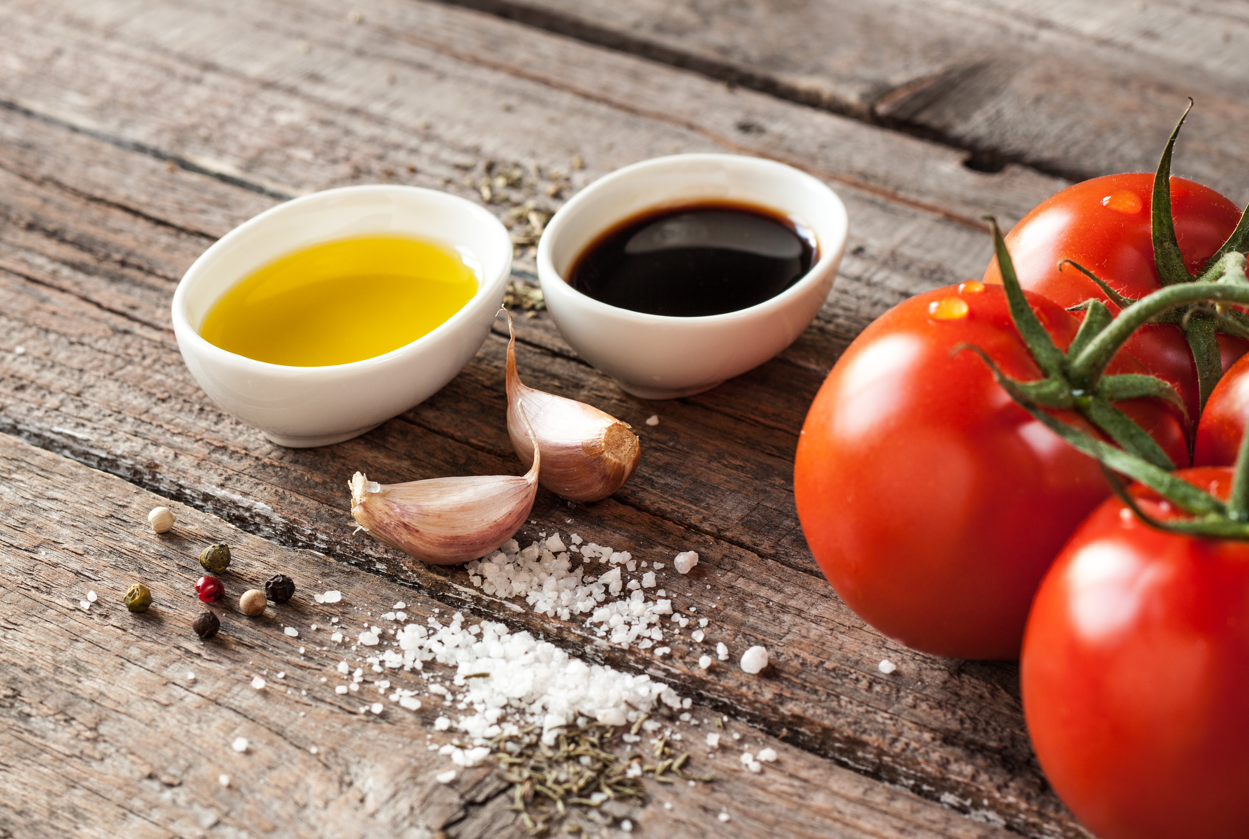 Two small white bowls, one containing oil and the other vinegar, next to a bunch of tomatoes, two garlic cloves, and seasonings on a wooden surface.