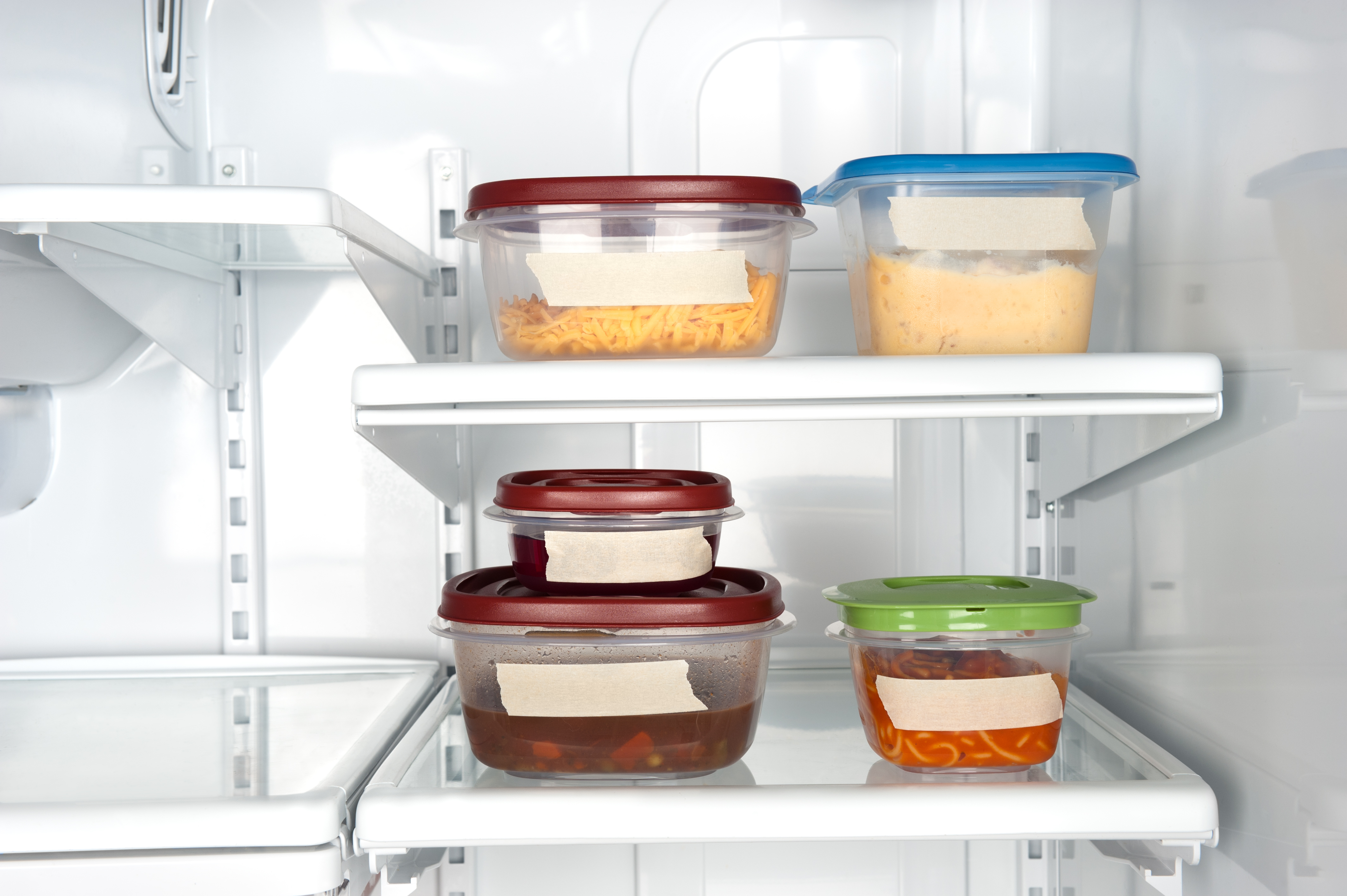 Five plastic containers filled with food with white labels in a refrigerator.