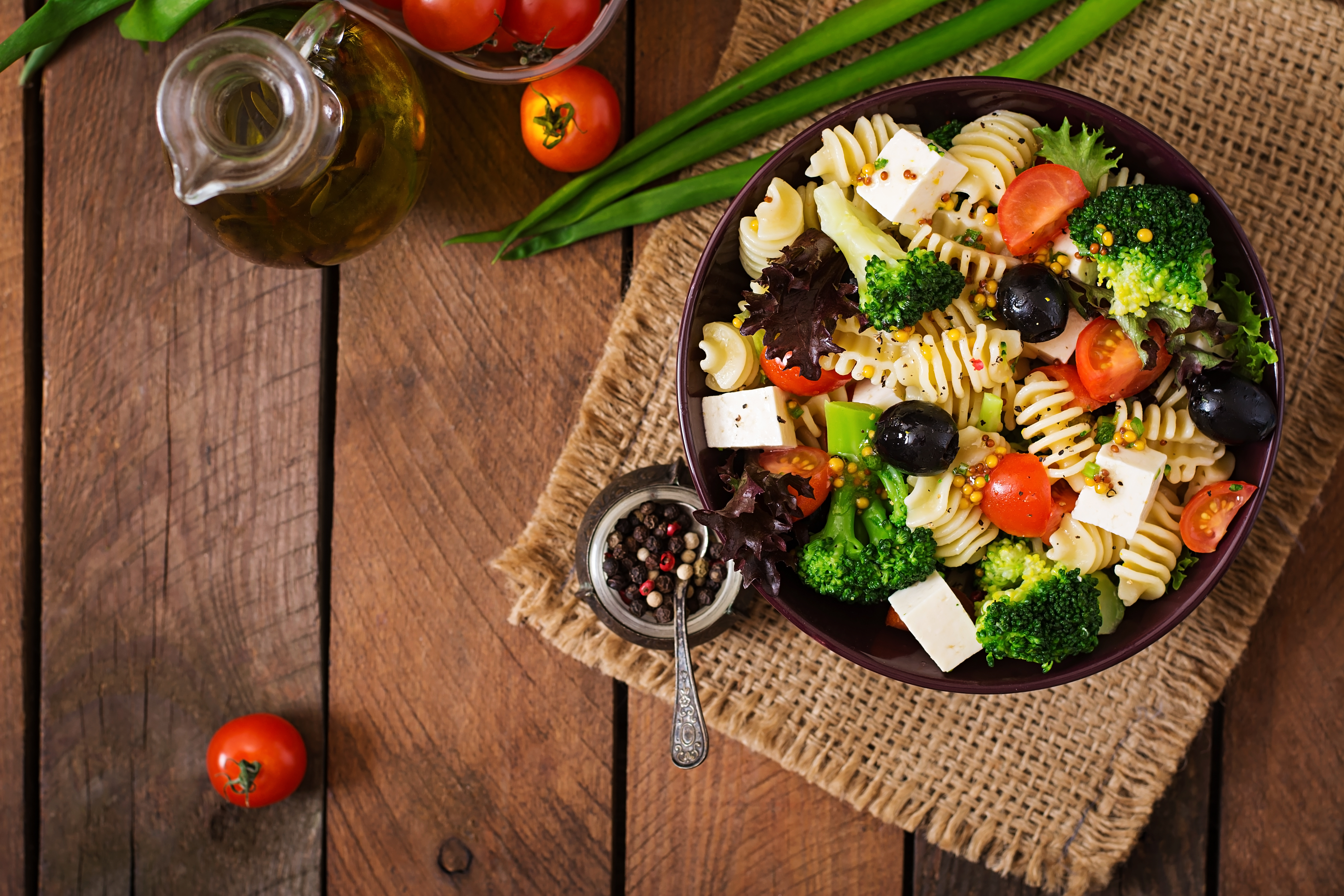 A bowl containing pasta, olives, tomatoes, broccoli, and cheese on top of a wooden surface.