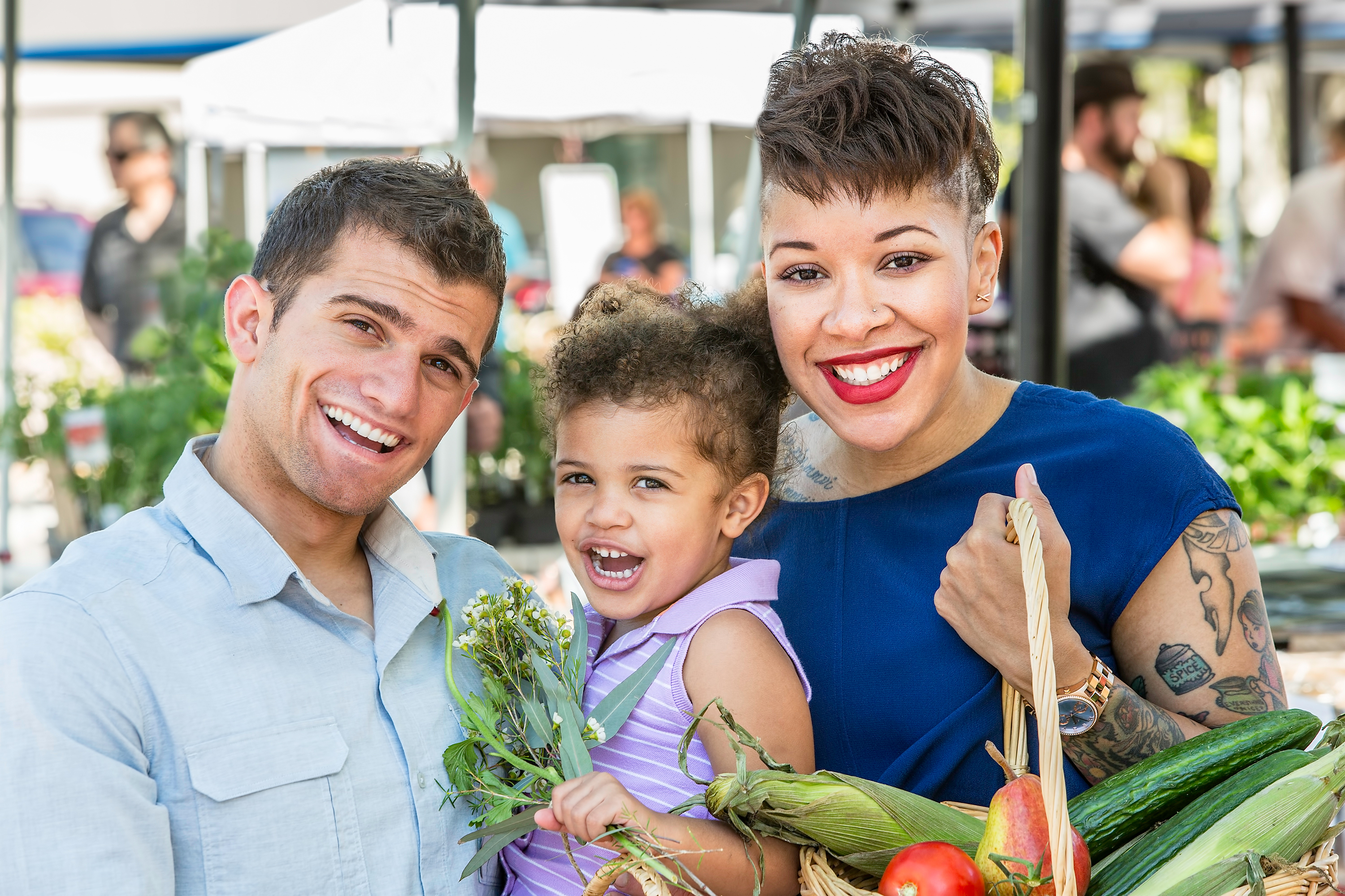 Parents holding a young child and a basket with fruits and vegetables while shopping at a farmers market.