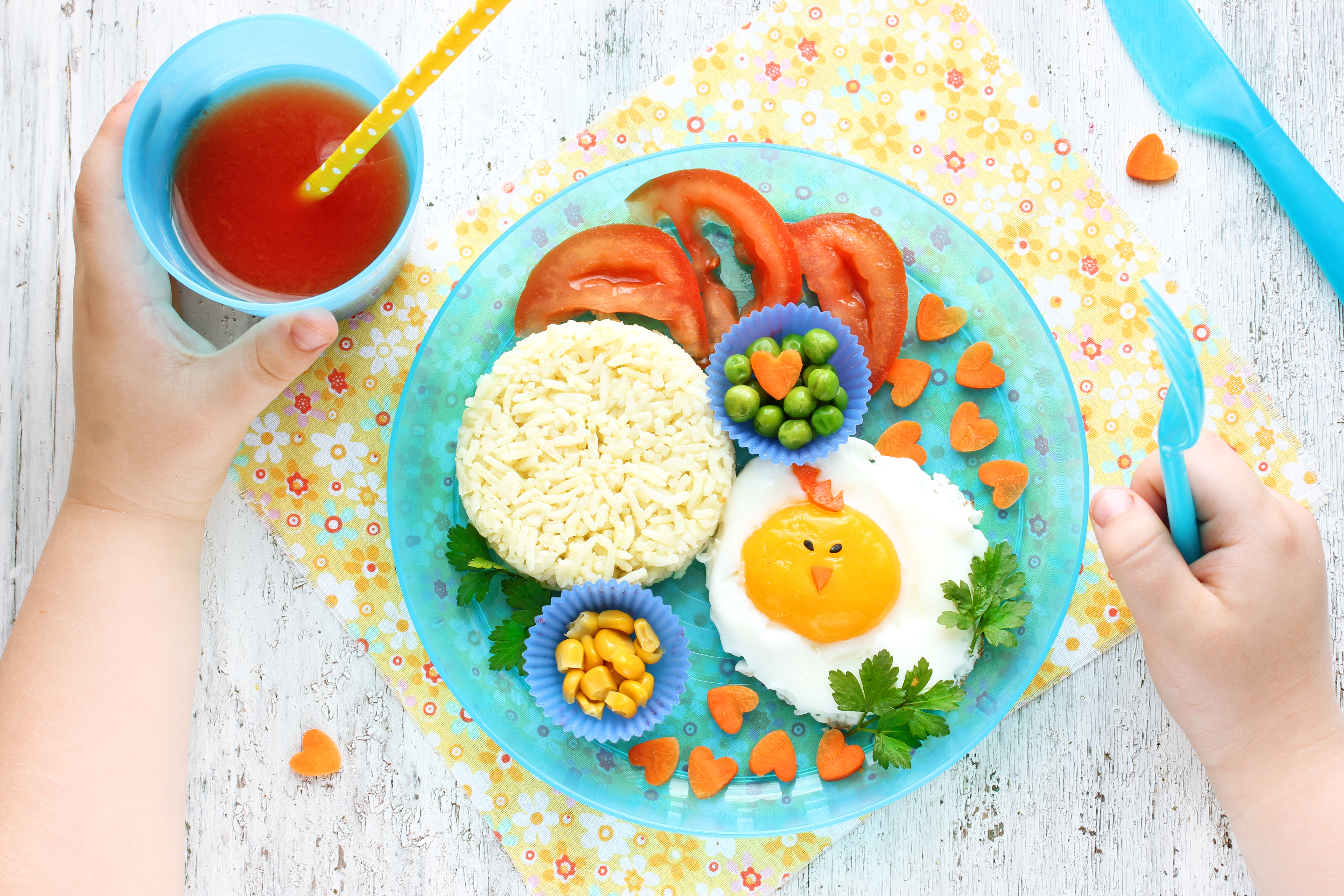A plate plate of rice, corn, peas, tomatoes, carrots cut into heart shapes, and a sunnyside-up egg. The plate is next to a childs hand holding a red beverage.