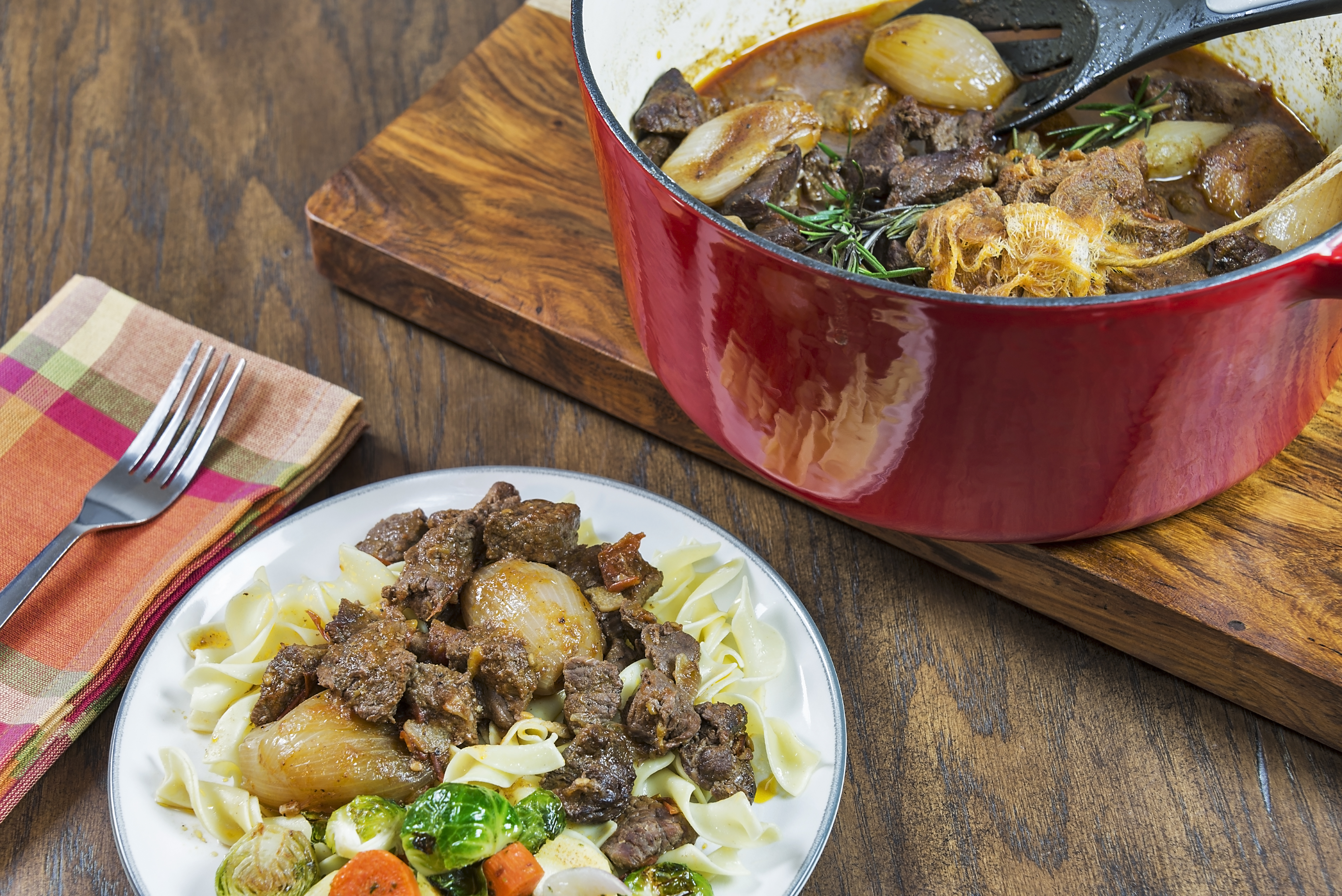 Red pot with vegetables and meat next to a plate with noodles topped with the meat and vegetables.