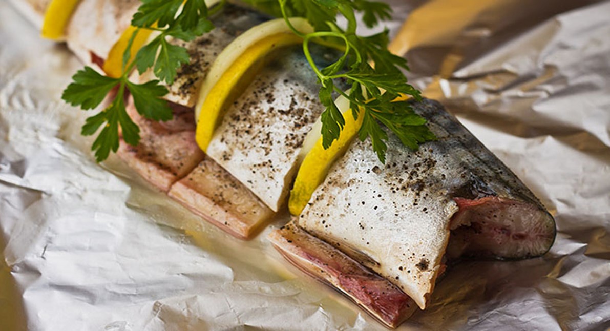 a foil cooked fish on a piece of foil topped with lemon slices and greens.