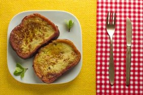 Two slices of French Toast on a plate, on w yellow table cloth, next to a red checkered napkin holding a fork and spoon.