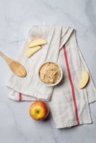 A dish of Peanut Butter Dip, a wooden spoon, and apple slices on a dish towel.