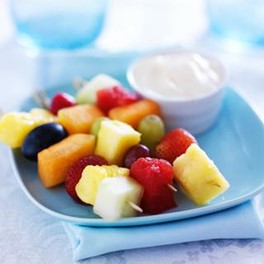 Three fruit kabobs on a plate with a dish of yogurt dip.