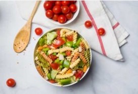 An image of pasta salad in a bowl next to a bowl of cherry tomatoes, a white napkin, and a wooden spoon.