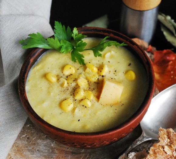 An image of potato, corn, and cheese soup in a red bowl with a sprig of parsley.