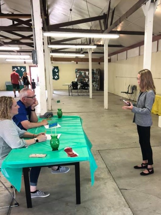 Female youth standing before a table with an adult male and adult female. 