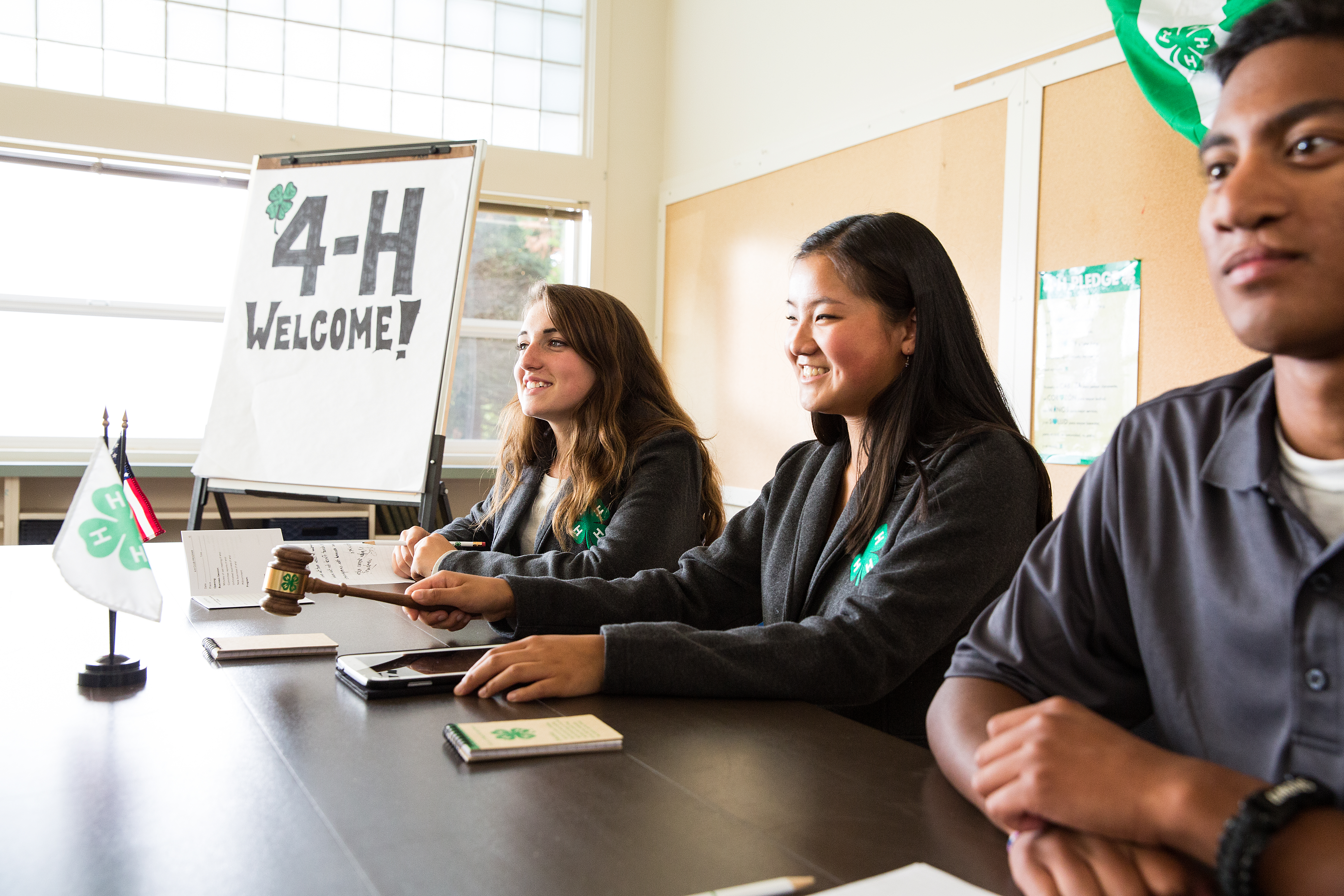 Left to right, two teenage girls and one teenage boy sitting at a head table in a classroom There is the American flag and 4-H flag poised on the table in front of the middle girl. She is also holding a gavel mid-air ready to tap the gavel stand in front of her. A 4-H Welcome! sign written on a flip chart on an eisel to the right of the girl on the far left.