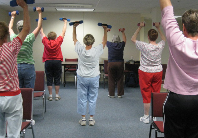 A group of women in an exercise class lift small weights over their heads.