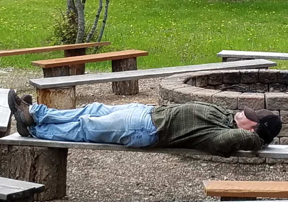 A man sleeps stretched out on a bench with a ball cap over his face.