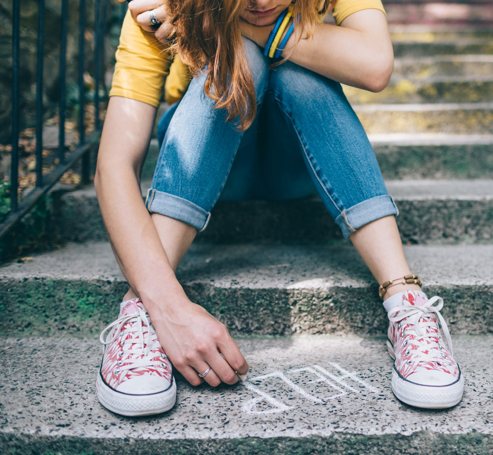 A adolescent girl sitting on concrete steps writes "help" in white chalk