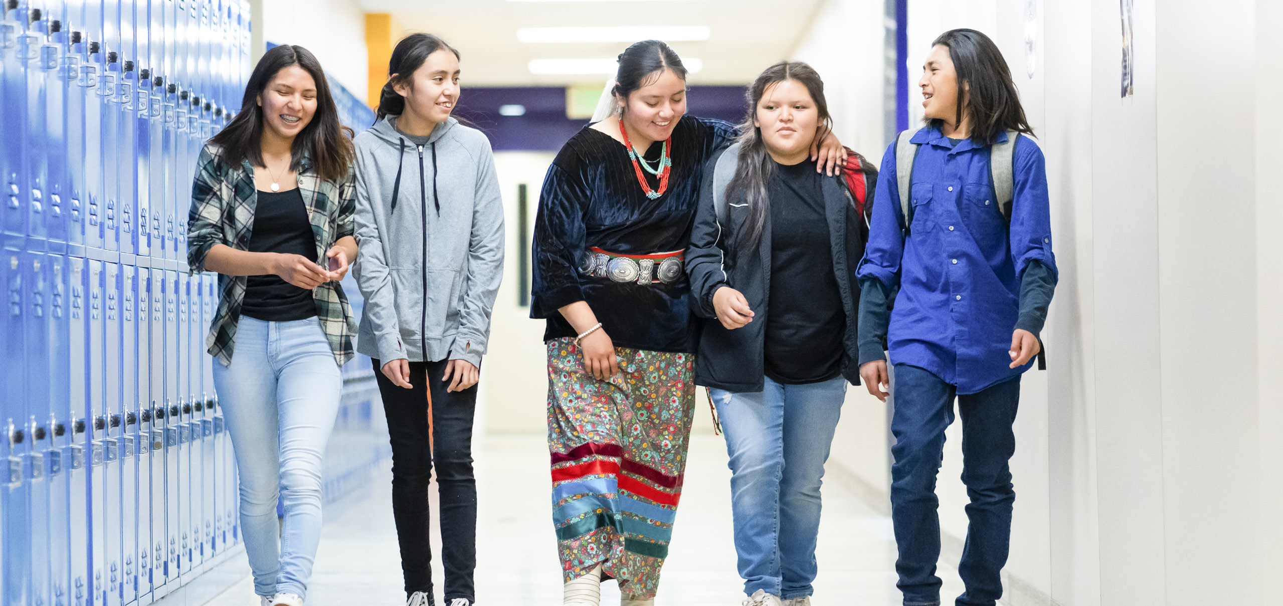 Five teens walk down a school hallway lined with lockers