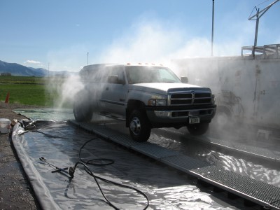 Silver pickup truck surrounded by mist from vehicle washing unit. Blue sky and green grass in the background.