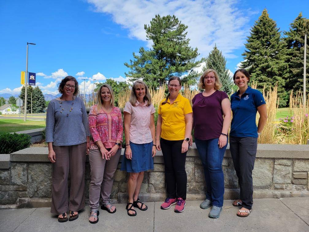 Six women standing in front of landscaping with blue sky and conifer trees in background