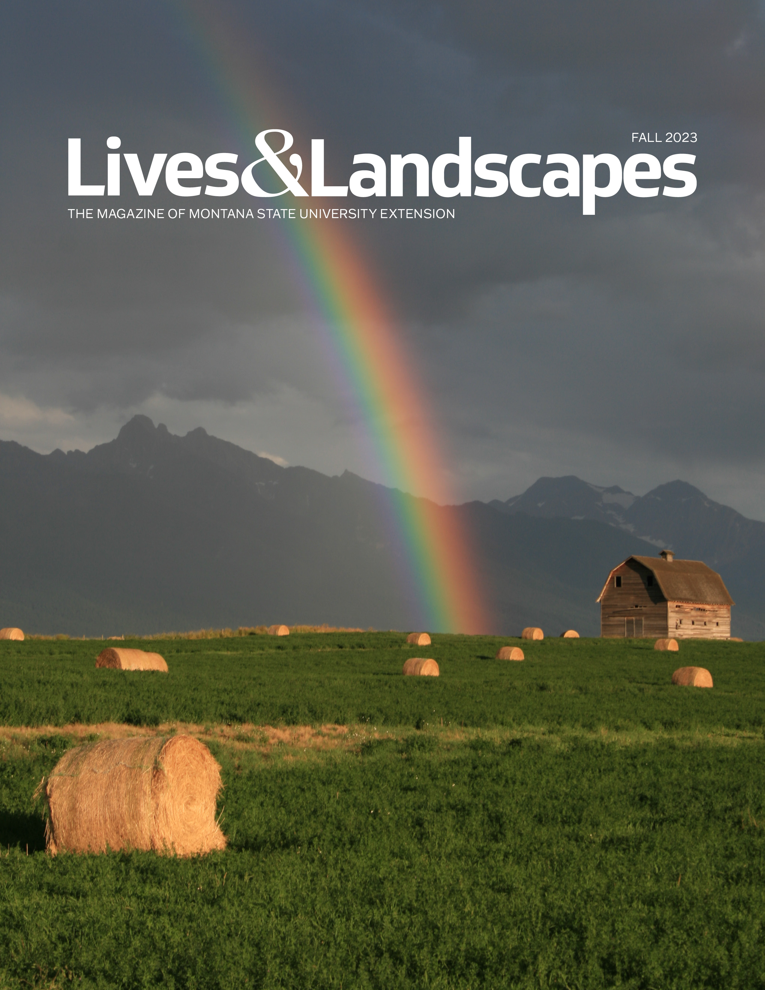 Cover image of Lives and Landscapes Fall 2023 features a striking photo of a rainbow coming out of a dark, stormy sky. and appearing to touch down in the middle of a green hay field dotted with golden hay bales. A brown barn sits off to one side. Barn and fields are lit up by the sun, while jagged peaks with bits of snow sit in shadow beneath the dark clouds in the background.