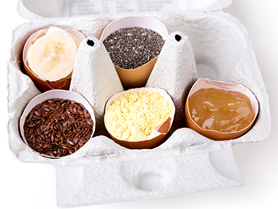 A carton for six eggs holds 5 half-shells. Each half contains a different egg substitute, such as chia seeds, flax seeds, banana, applesauce, and egg replacer powder.