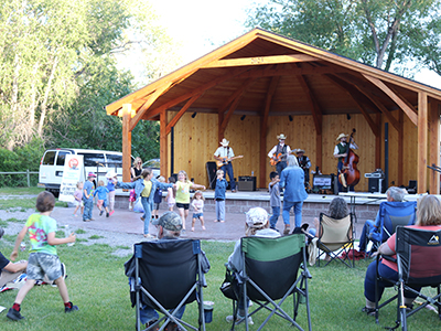 Spectators in camp chairs watch and children dance in front of a wooden bandstand with a 4-person band playing inside