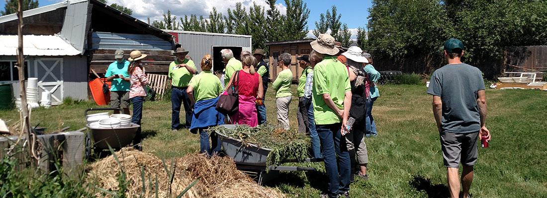 Group of people standing in a grassy area on a farm, listening to a speaker