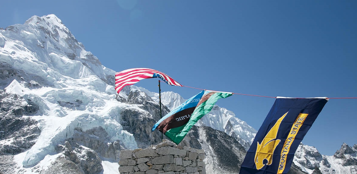 American flag, Montana State flag, and undescript flag hanging from a line with scenic, snow-covered mountain in background