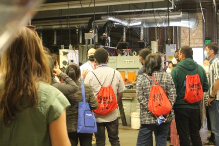 Photo of the backs of several people standing in a garage (autoshop class).They are all wearing Hawkers backpacks and look adorable. They appear very engaged with the speaker of the group.