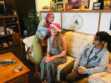 Two young women put tradition headwear on another woman while sitting on a couch.