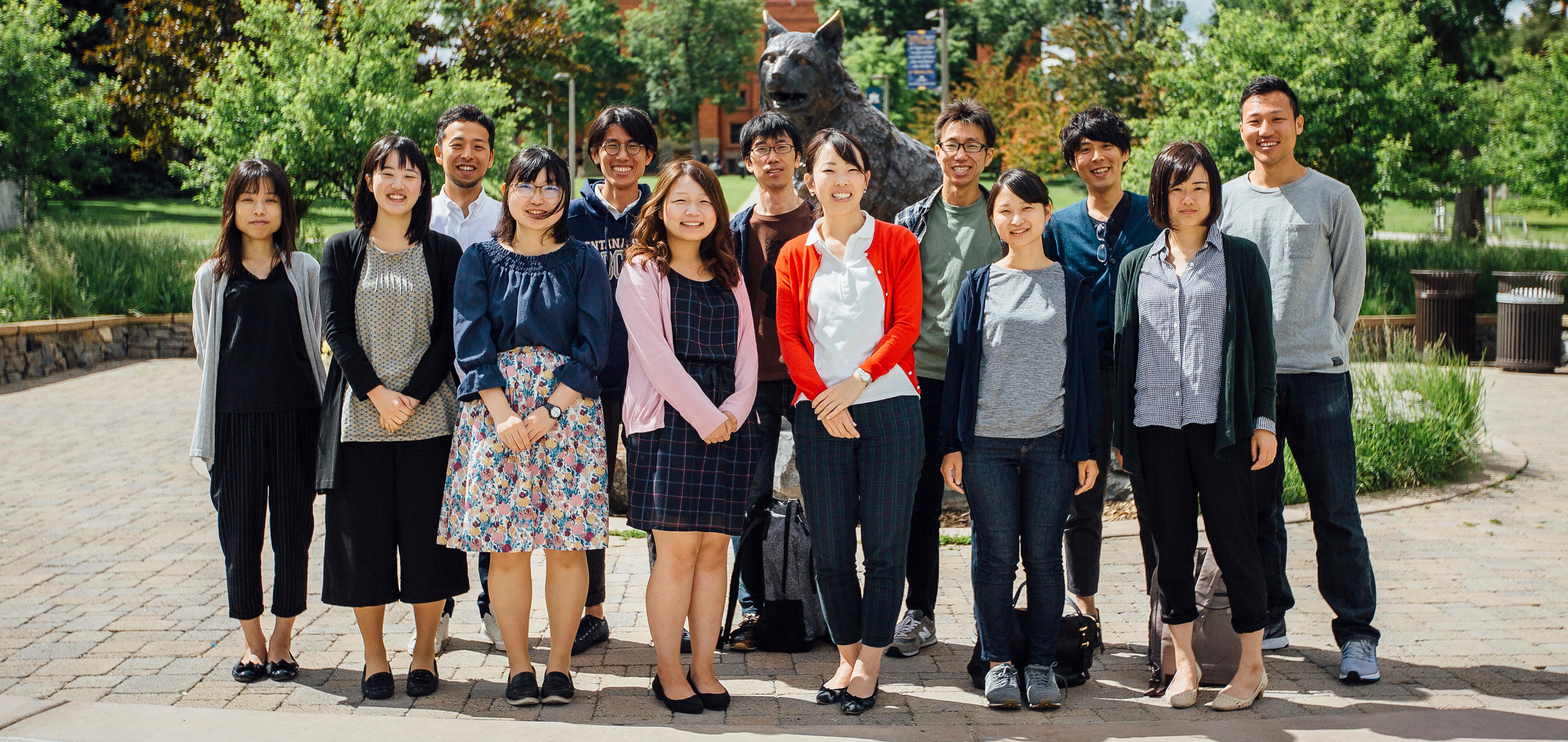 A group of Japanese visitors pose in front of a statue of a bobcat on MSU campus. It looks lovely outside and is very green out.