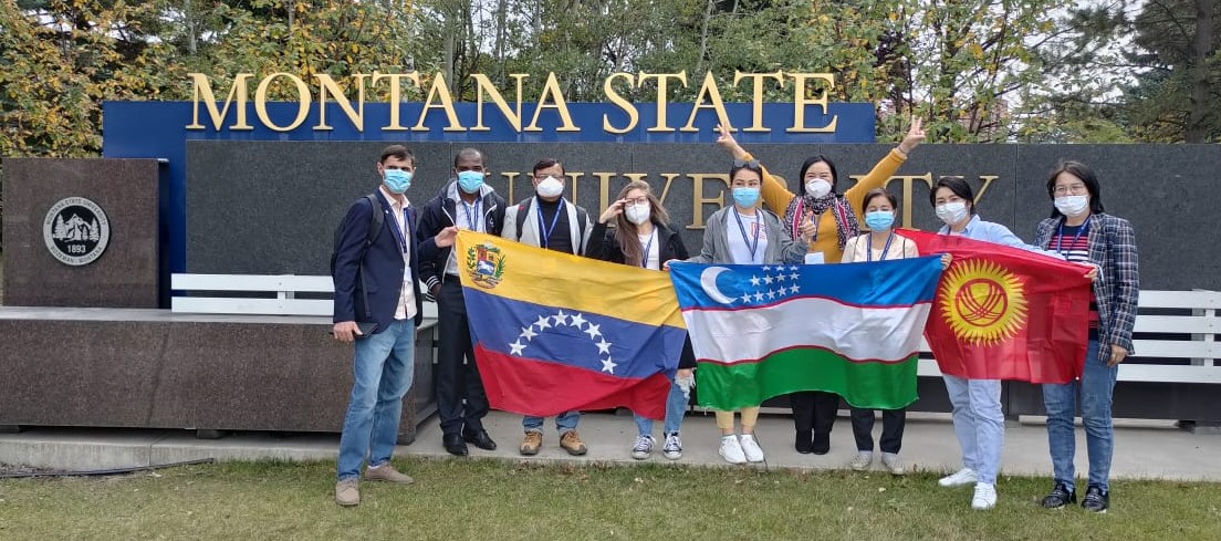 9 Fulbright TEA Fellows stand in front of an MSU sign holding their home country's' flags up.