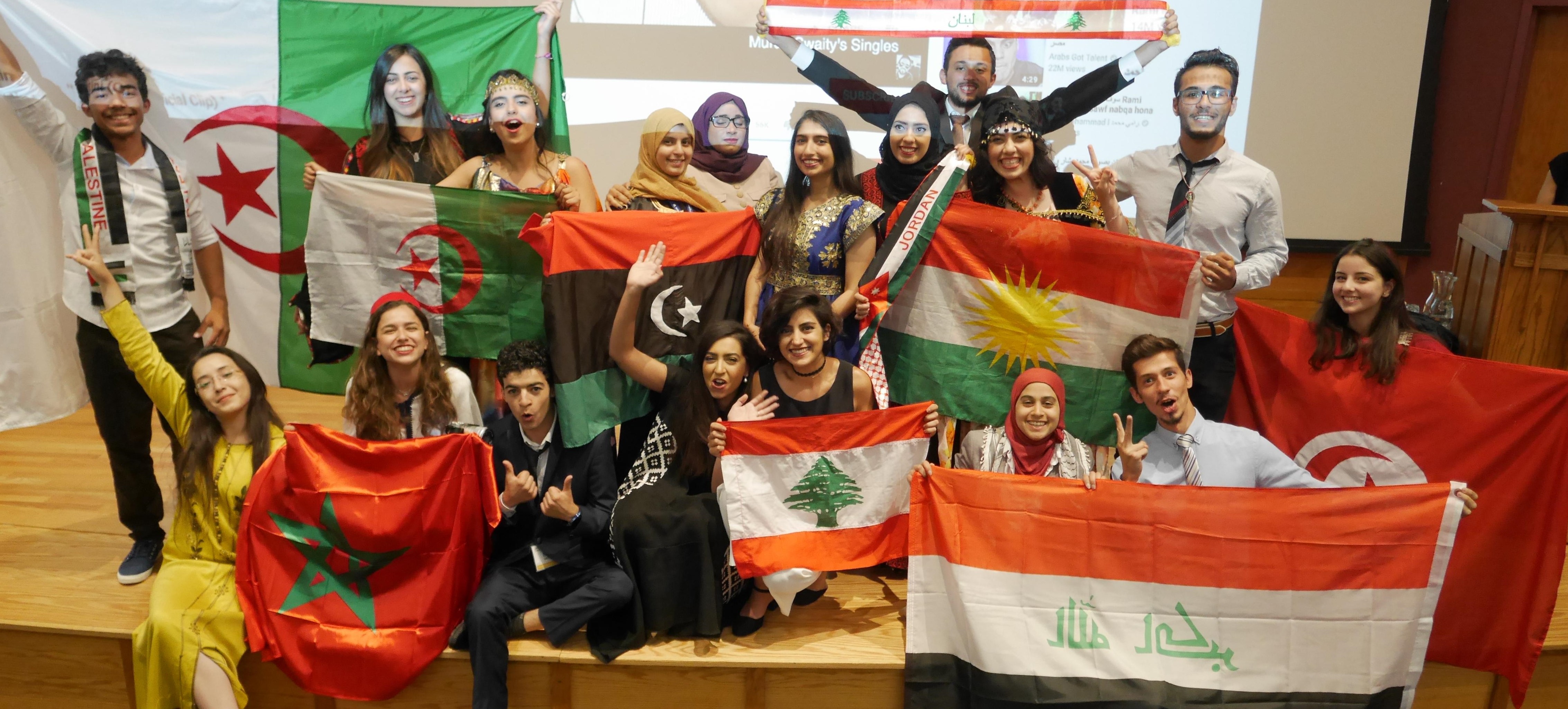 Young students stabd on a stage in traditional Middle Eastern and North African wear, looking psyched and holding flags.